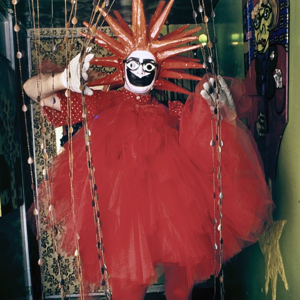 El artista de performance Leigh Bowery posa en su departamento vistiendo un traje rojo con un voluminoso tutú de tul, guantes blancos brillantes, una máscara de payaso grotesca y un tocado de picos rojos tipo sol radiante, detrás de una cortina de cuentas.