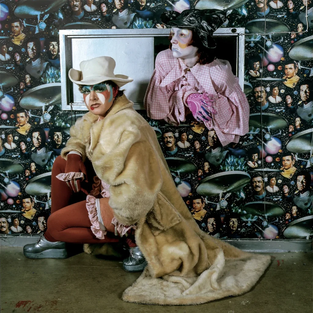 El artista Leigh Bowery con abrigo de piel sintética y sombrero blanco posa junto a Trojan (Guy Furrow), quien asoma por una ventanilla con blusa a cuadros rosa frente a una pared con papel tapiz de Star Trek en Londres, 1984.