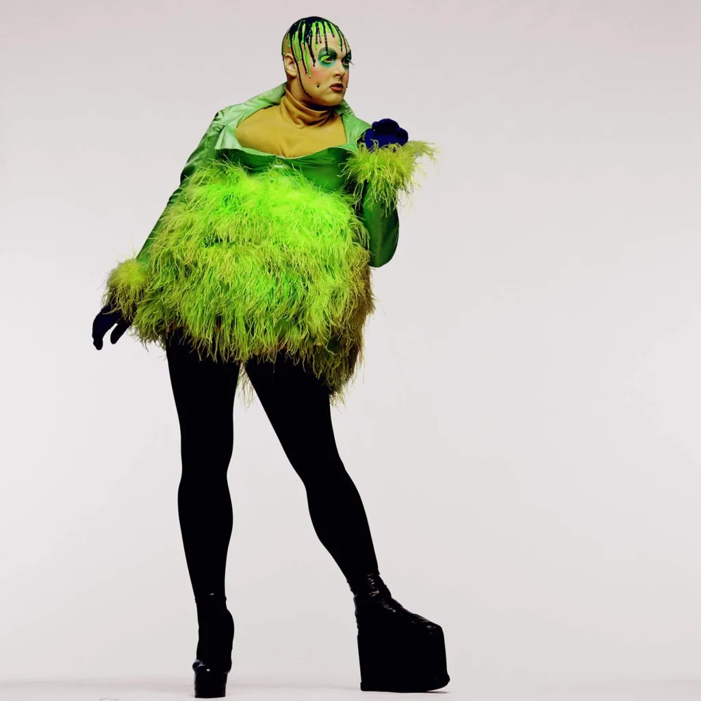 El artista de performance Leigh Bowery posa de cuerpo entero usando un vestido verde menta con una voluminosa falda de plumas verdes, guantes negros, medias negras y botas de plataforma gigantes, fotografiado por Fergus Greer en 1991.