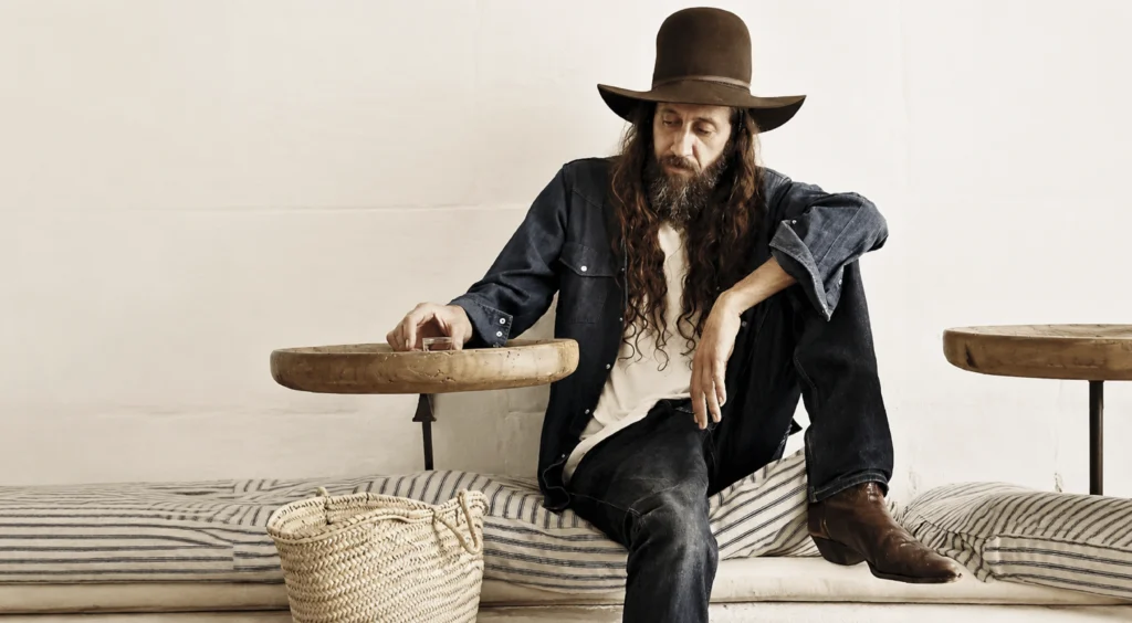 El diseñador de moda español Miguel Adrover con sombrero vaquero de fieltro marrón y camisa vaquera azul, sentado en un banco de su bar Es Jaç en Mallorca. Retrato editorial de Germán Sáiz para El País S Moda.