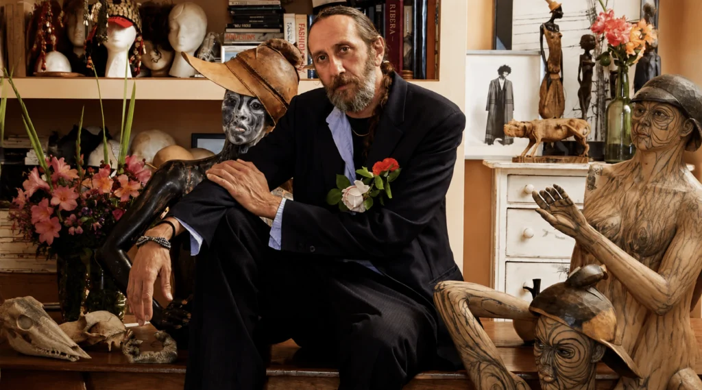 El diseñador de moda español Miguel Adrover con traje oscuro y flores en la solapa, sentado en su taller artístico de Calonge en Mallorca junto a maniquíes y figuras esculpidas. Retrato oficial para Vogue España.