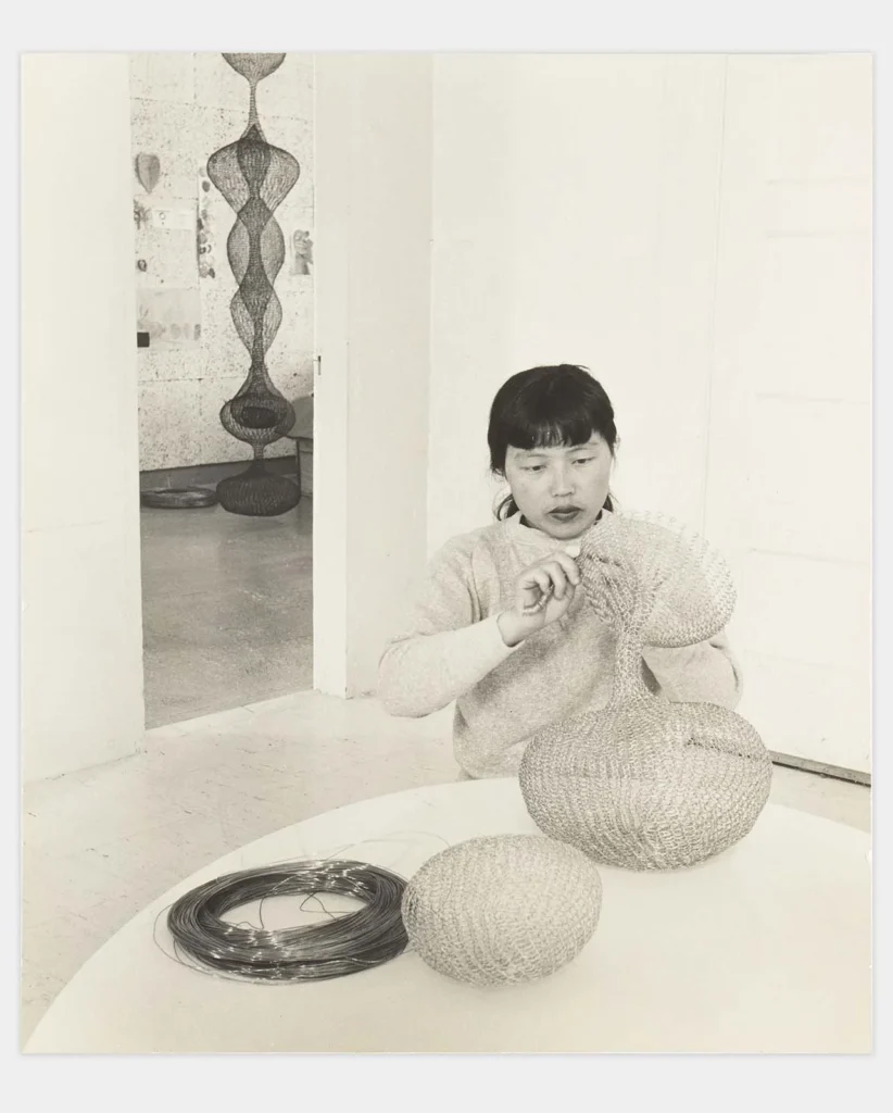 Ruth Asawa trabajando en su taller con rollos de alambre y esculturas terminadas al fondo, ejemplificando su técnica de dibujo tridimensional y transparencia visual.