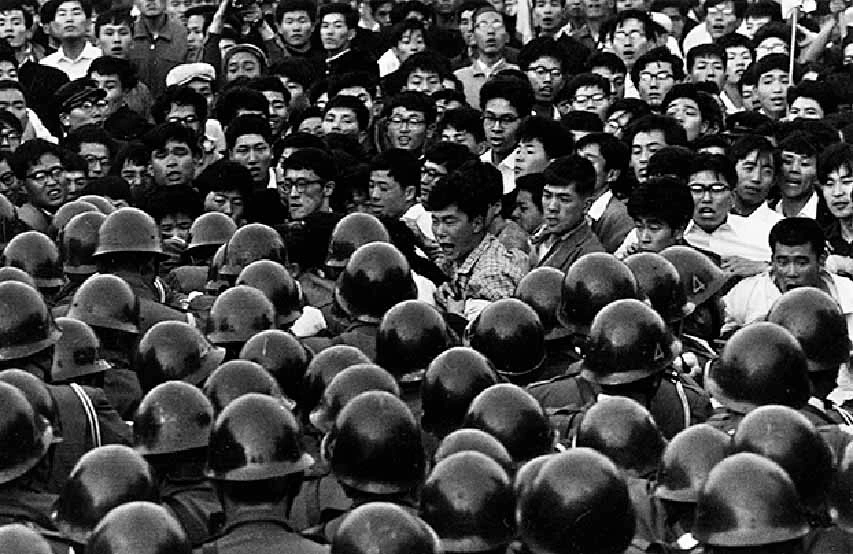 Fotografía documental en blanco y negro de Takuma Nakahira en 1966, que muestra a un joven manifestante gritando en medio de una multitud civil confrontada por un mar de policías con cascos militares oscuros en Tokio.