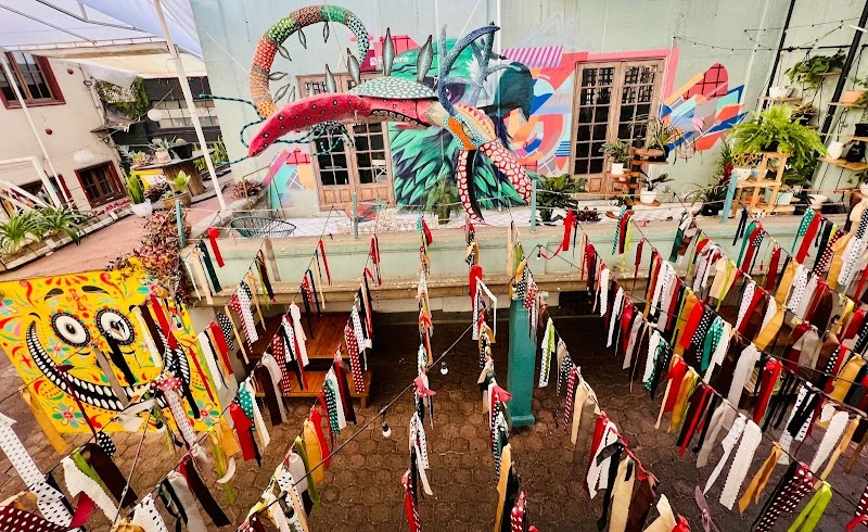 Patio cultural decorado con un alebrije colgante, murales coloridos y tiras de listones en el Tianguis de la Bola en Coyoacán, CDMX.