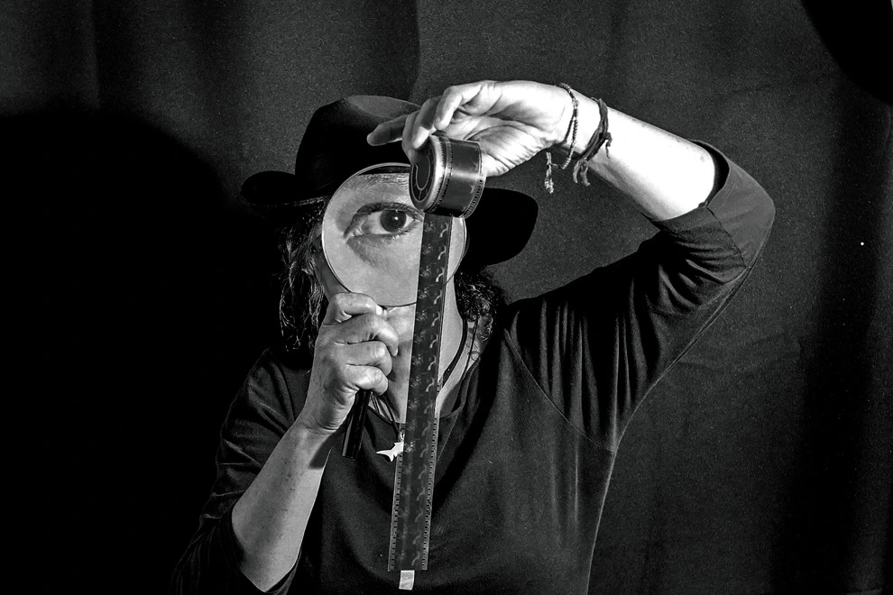 Retrato conceptual en blanco y negro de Ximena Cuevas usando sombrero, sosteniendo una lupa que amplifica su ojo izquierdo y desenrollando una tira de película cinematográfica.