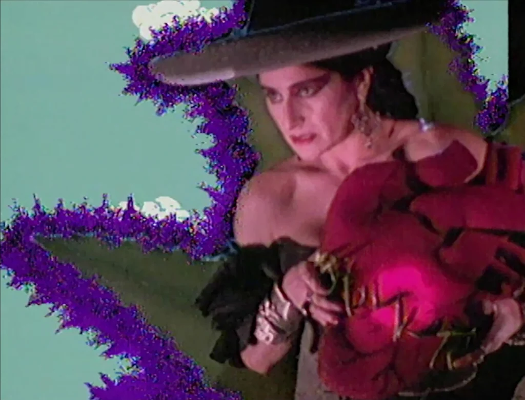Fotograma de videoarte de Ximena Cuevas usando un sombrero de ala ancha y cargando un gran corazón sangrante artificial con un fondo editado en tonos verdes y morados.