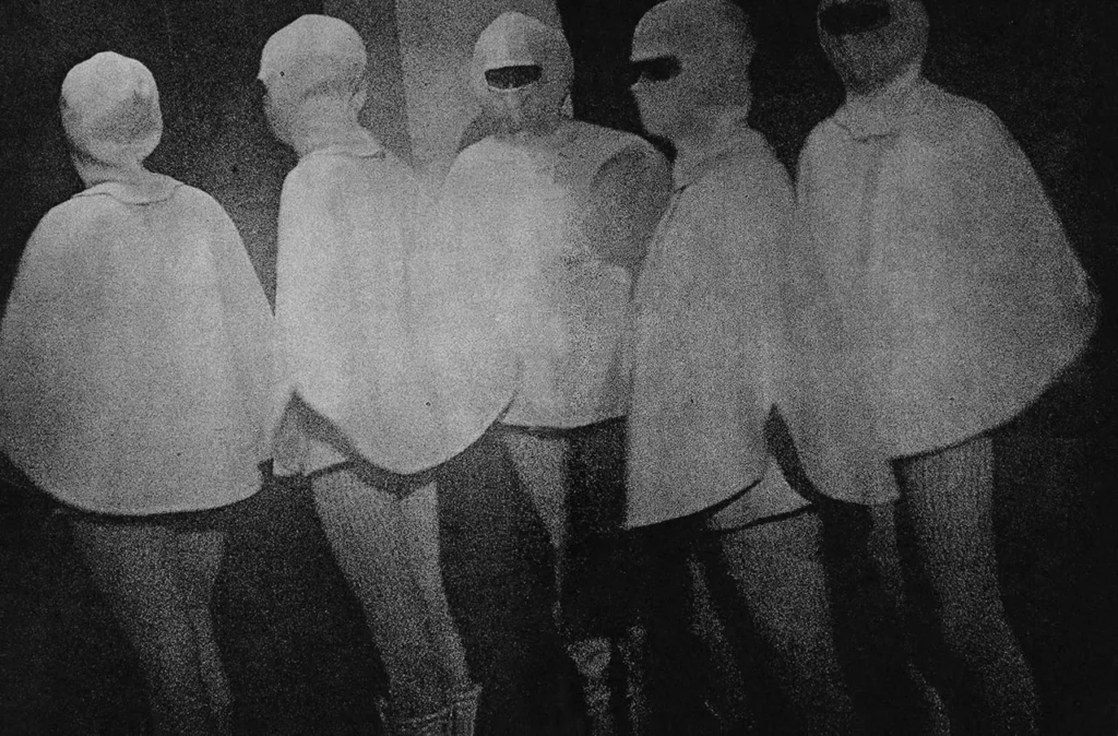 Fotografía analógica de Yutaka Takanashi de 1969 en estilo are bure bokeh, que muestra un grupo de cinco personas alineadas de espaldas y de perfil vistiendo capas blancas idénticas y capuchas en una atmósfera nocturna y granulada.
