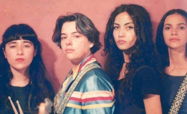 Fotografía en plano medio y a color de las cuatro integrantes originales de la banda mexicana Las Ultrasónicas a mediados de los años 90. De izquierda a derecha aparecen la baterista Jenny Bombo sosteniendo unas baquetas, la tecladista Tere Farfisa con una chaqueta satinada azul con franjas rojas, y las integrantes Jessy Bulbo y Suzy Vox de perfil parcial. Posan con semblante serio frente a una pared texturizada en tonos rojizos.