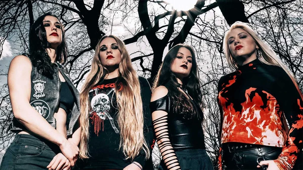 Fotografía promocional en plano medio de las cuatro integrantes de la banda de metal Nervosa posando al aire libre bajo árboles sin hojas. De izquierda a derecha: la bajista Hel Pyre con chaleco de parches de bandas; la guitarrista y vocalista Prika Amaral con playera negra de calavera; la guitarrista Helena Kotina con blusa de mangas recortadas; y la baterista Michaela Naydenova con una blusa de estampado de fuego rojo y negro. Todas miran con semblante serio y desafiante hacia la cámara.