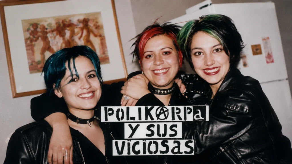 Fotografía a color de plano medio de las tres integrantes de la banda colombiana de punk Polikarpa y Sus Viciosas, abrazadas y sonriendo a la cámara. Tienen peinados cortos con tintes de colores (azul, rojo y verde) y visten chaquetas de cuero negro, gargantillas con picos metálicos (chokers) y maquillaje oscuro. En la parte inferior, superpuesto sobre la imagen, aparece un cartel con letras recortadas estilo fanzine que deletrea el nombre de la banda 
