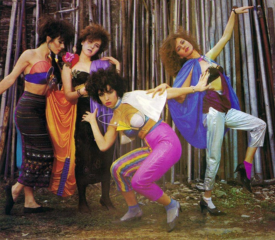 Fotografía a color de las cuatro integrantes de la banda argentina Viuda e Hijas de Roque Enroll posando al aire libre frente a una estructura de tubos metálicos. Visten ropa llamativa de los años 80: de izquierda a derecha, una integrante con falda larga oscura y top morado; otra con vestido negro y detalles naranjas; al centro, una agachada con pantalones rosa brillante y corsé futurista; y a la derecha, una integrante con pantalones plateados y capa azul translúcida en una pose teatral.