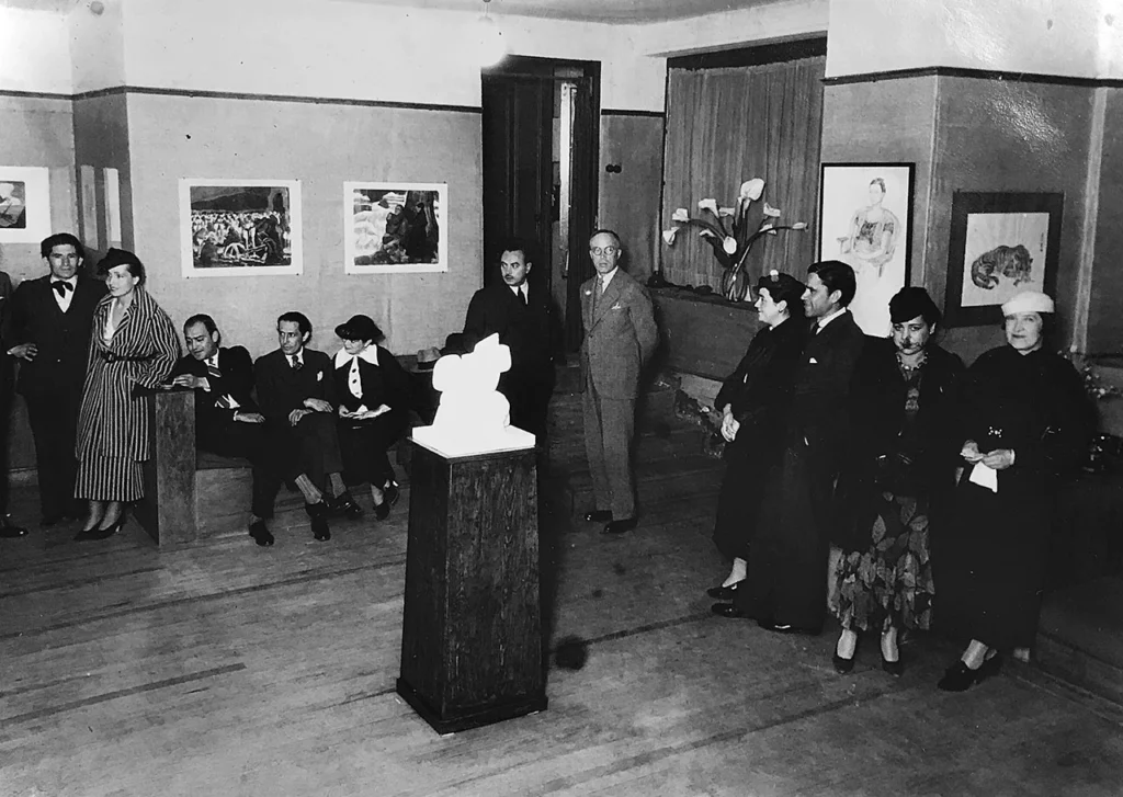 Fotografía antigua en blanco y negro de una inauguración artística en la Galería de Arte Mexicano, con asistentes elegantemente vestidos, cuadros colgados en los muros y una escultura abstracta sobre un pedestal de madera.