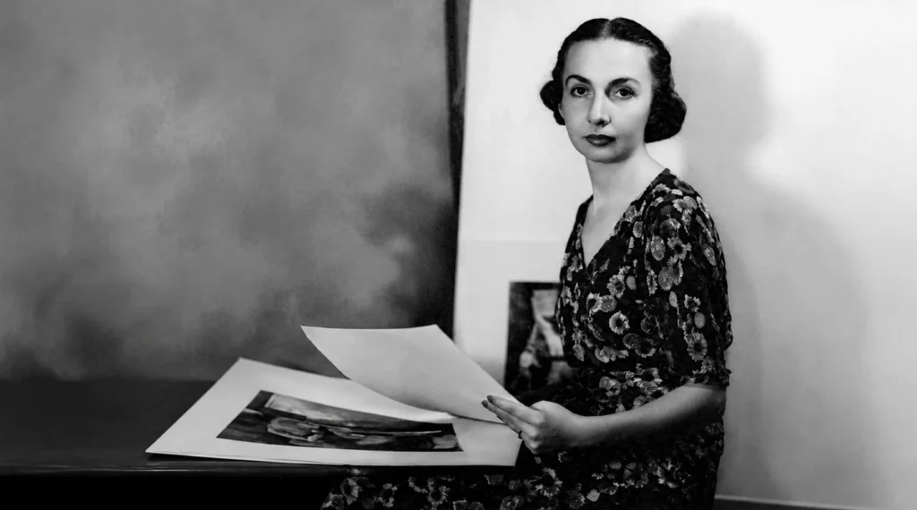 Retrato en blanco y negro de una joven Inés Amor sentada junto a una mesa, sosteniendo y revisando grabados o documentos de arte en papel.