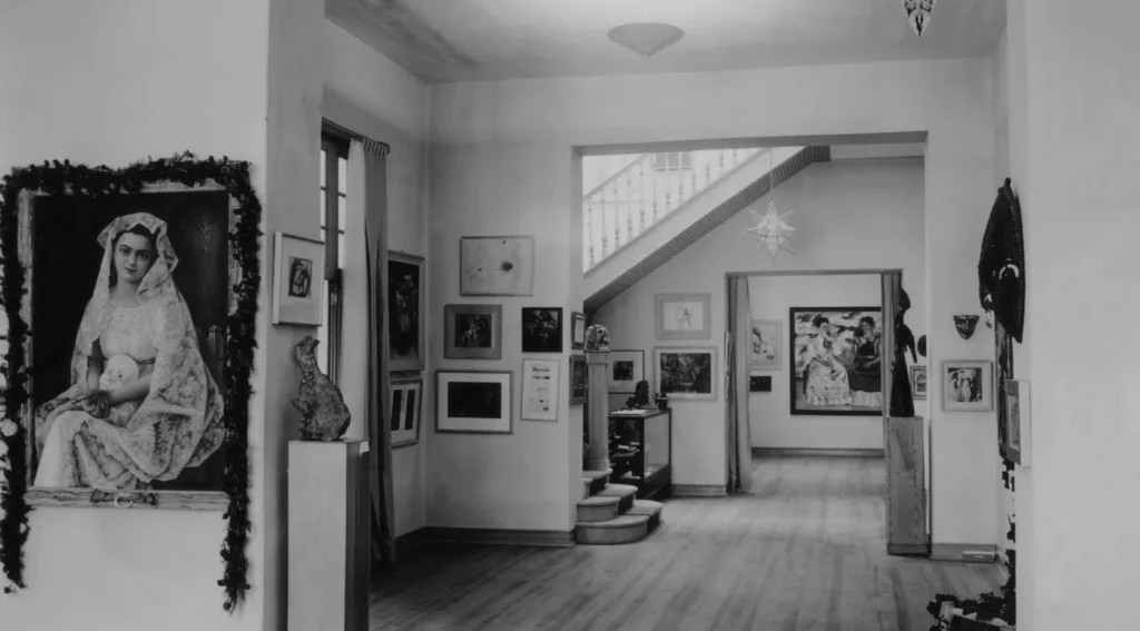 Fotografía en blanco y negro del interior de la Galería de Arte Mexicano, mostrando salones comunicados, una escalera al fondo, múltiples pinturas colgadas en los muros y esculturas en pedestales.