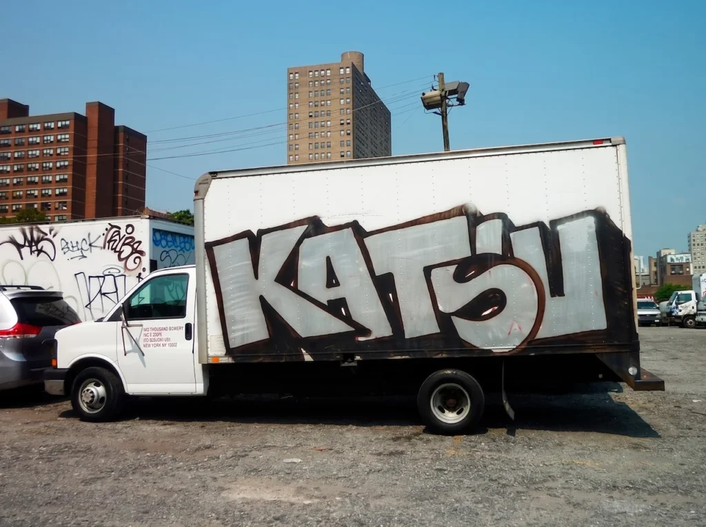 Camión de carga blanco con un tag monumental de KATSU en color plata y negro, estacionado en un entorno urbano de Nueva York, ilustrando la escala industrial de su obra analógica.