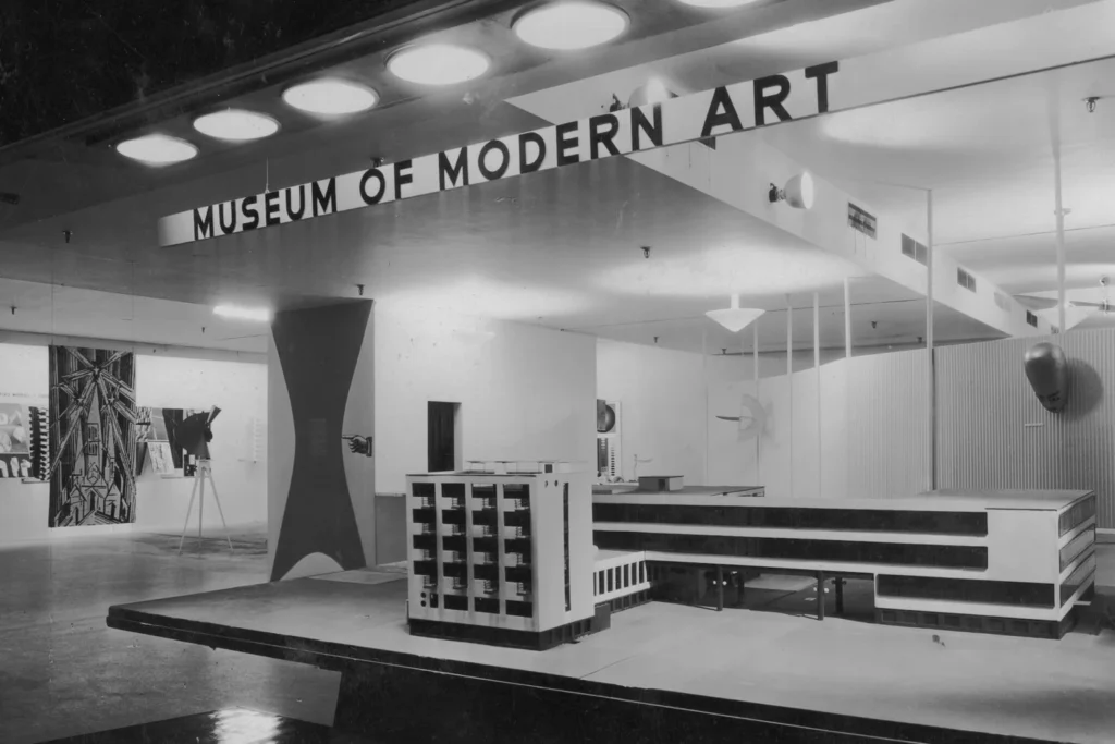 Fotografía antigua en blanco y negro del interior de una sala del Museum of Modern Art de Nueva York, mostrando maquetas arquitectónicas y paneles informativos expuestos.