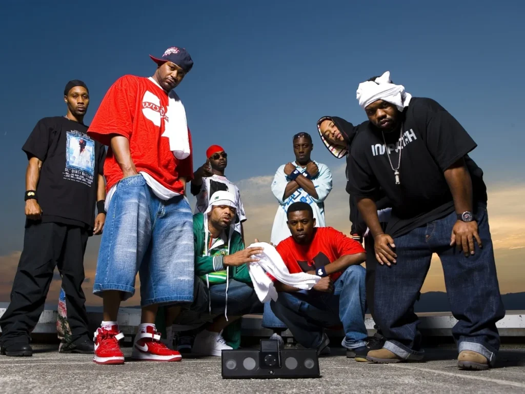 Fotografía a color del grupo Wu-Tang Clan posando en una azotea durante el atardecer. Los integrantes visten ropa urbana contemporánea con playeras de marcas icónicas y sudaderas. En el centro del grupo se aprecia un reproductor de música portátil, simbolizando el impacto del colectivo en la tecnología y la difusión del hip hop moderno.