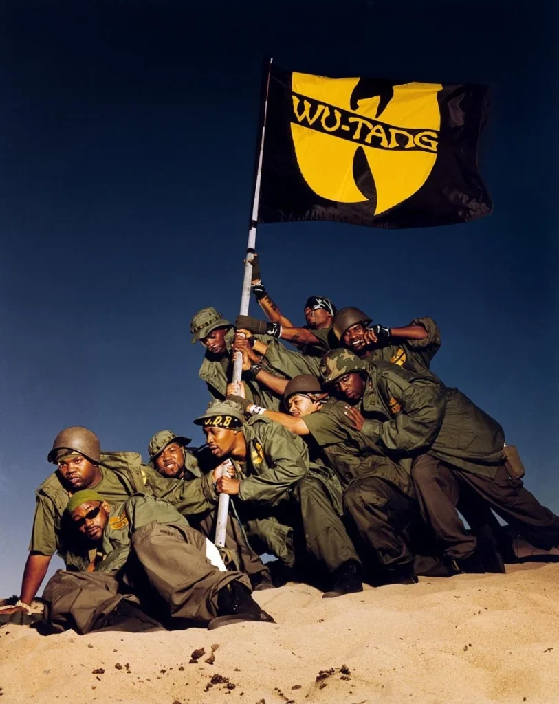 Fotografía del grupo Wu-Tang Clan recreando la icónica pose de los soldados en Iwo Jima, levantando una bandera negra con el logo amarillo del Wu en un paisaje desértico. Los integrantes visten uniformes militares de color verde oliva, simbolizando la unidad y la conquista del grupo en la industria del hip hop.