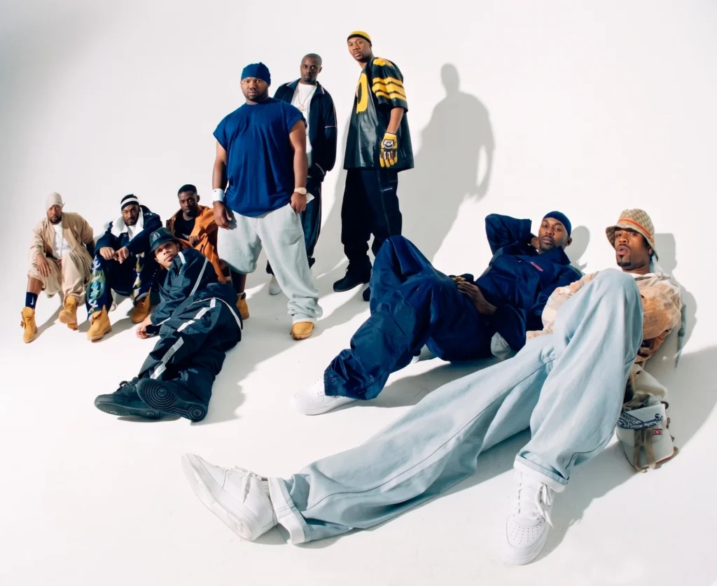 Fotografía en plano contrapicado de los integrantes de Wu-Tang Clan en un estudio de fondo blanco, luciendo ropa 'baggy' característica de los años 90 como sudaderas, gorros y calzado deportivo. La imagen captura la estética visual del hip hop de la Costa Este y la identidad colectiva del grupo liderado por RZA.