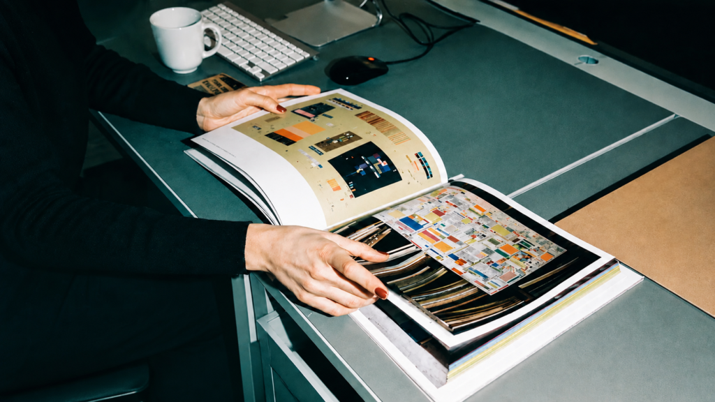 Primer plano de dos personas revisando un libro de arte y diseño editorial abierto sobre un escritorio gris moderno. En el fondo desenfocado se aprecian un teclado de computadora blanco Apple, un ratón negro y una taza blanca de café.
