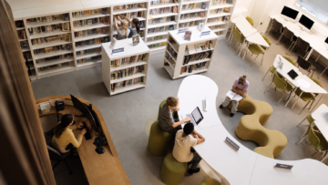 Las mejores bibliotecas virtuales para leer y descargar libros gratis Vista aérea de una biblioteca escolar moderna con estudiantes usando laptops y mesas curvas.