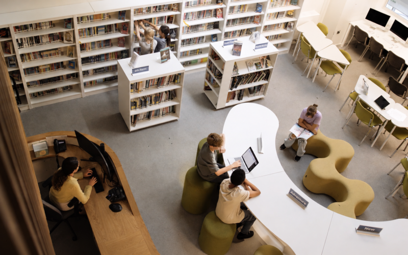 Vista aérea de una biblioteca escolar moderna con estudiantes usando laptops y mesas curvas.