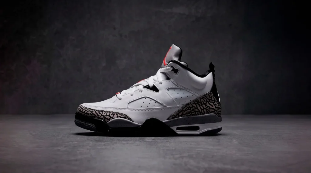 Tenis Jordan de perfil deportivo en color blanco con detalles en negro, gris y un patrón texturizado grisáceo en la punta y el talón, fotografiado de perfil sobre un fondo de cemento gris.