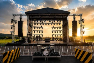 La infraestructura invisible de los festivales de música Escenario principal vacío de un festival de música al aire libre con torres de luces, altavoces line array suspendidos, batería armada en primer plano y atardecer de fondo.