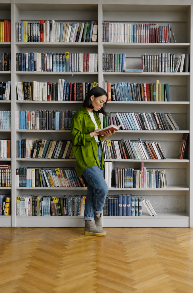 Mujer joven con lentes leyendo un libro frente a una estantería llena en una biblioteca.
