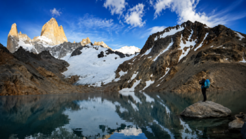 Senderista con mochila azul sobre una roca contemplando el reflejo del Monte Fitz Roy en la Laguna de los Tres, Parque Nacional Los Glaciares, Patagonia Argentina.