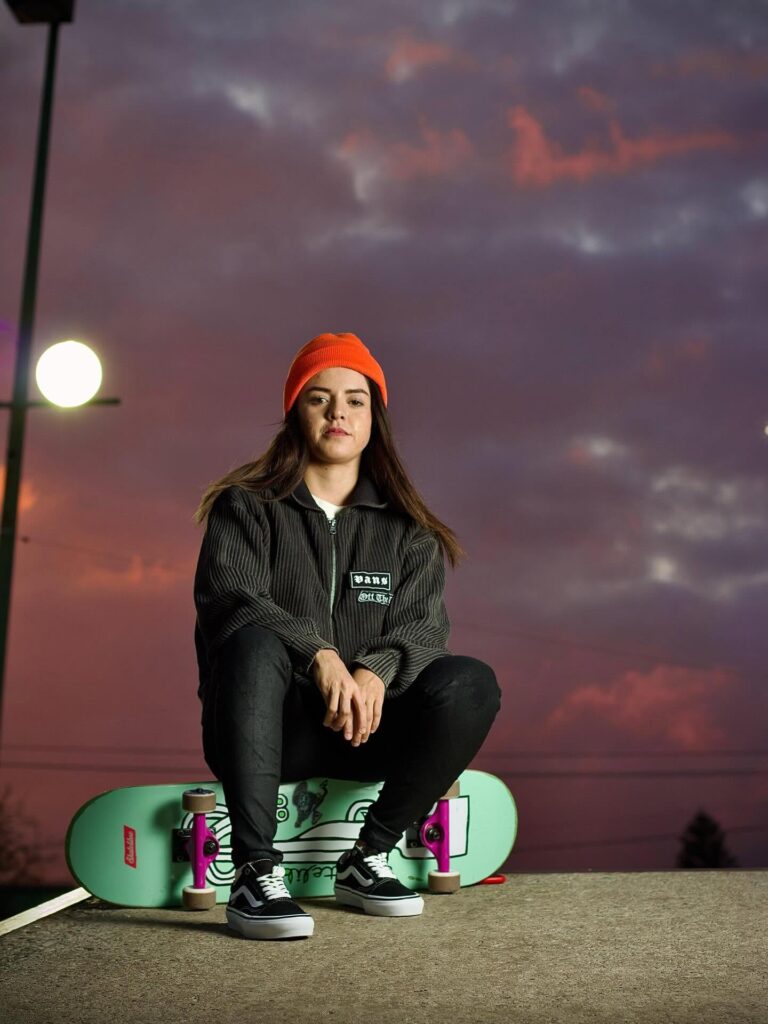 Retrato de una skater mexicana con gorro naranja y chaqueta Vans sentada junto a su patineta verde frente a un cielo crepuscular.