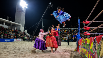 Cuatro cholitas luchadoras bolivianas en un ring de lucha libre sobre la arena de playa en Arena Cavancha, Iquique, Chile. Una de las luchadoras ejecuta un salto aéreo con su pollera tradicional celeste extendida mientras sus compañeras esperan abajo para recibir el impacto, frente a un público masivo por la noche.