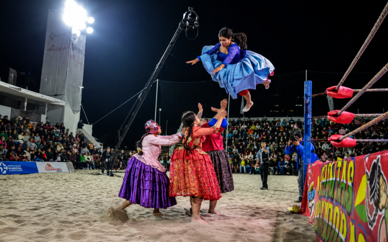 Cuatro cholitas luchadoras bolivianas en un ring de lucha libre sobre la arena de playa en Arena Cavancha, Iquique, Chile. Una de las luchadoras ejecuta un salto aéreo con su pollera tradicional celeste extendida mientras sus compañeras esperan abajo para recibir el impacto, frente a un público masivo por la noche.