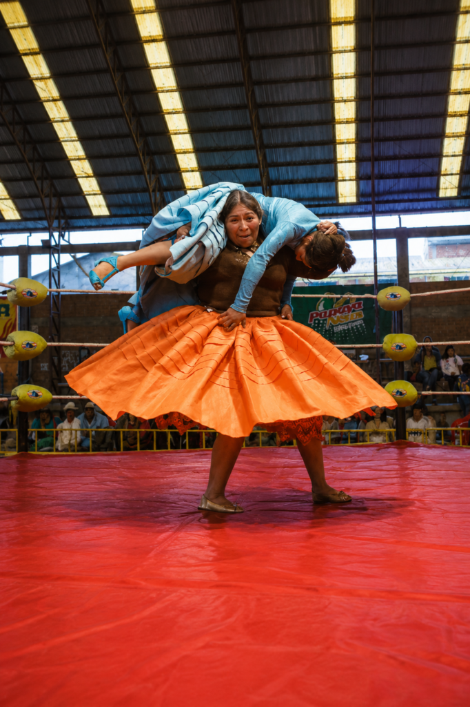 Una cholita luchadora boliviana con una pollera naranja brillante carga sobre sus hombros a otra luchadora vestida de azul celeste. Ambas se encuentran en el centro de un ring de lucha libre con lona roja dentro de un coliseo techado en El Alto, Bolivia, frente al público.