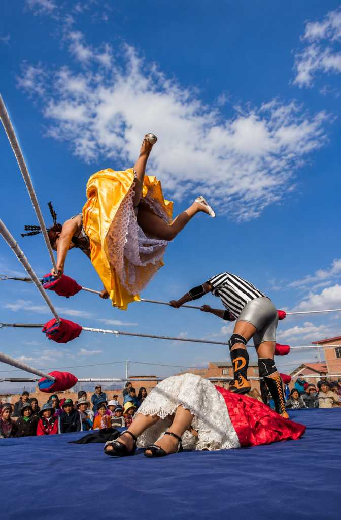Una cholita luchadora boliviana salta con acrobacia aérea desde las cuerdas usando una pollera amarilla y enaguas blancas. Abajo en el ring azul, un árbitro de camiseta de rayas observa a otra luchadora con pollera roja tendida en la lona, durante un show masivo al aire libre bajo un cielo azul con nubes.
