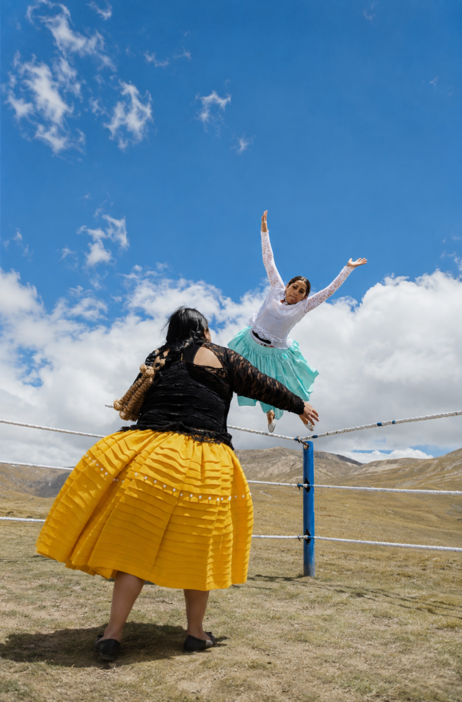 Una cholita luchadora boliviana salta por los aires con los brazos abiertos en un ring al aire libre, vistiendo una pollera turquesa y blusa blanca. En primer plano, otra luchadora de espaldas con pollera amarilla y blusa negra observa el salto en la altura andina de El Alto, Bolivia, bajo un cielo azul con nubes blancas.