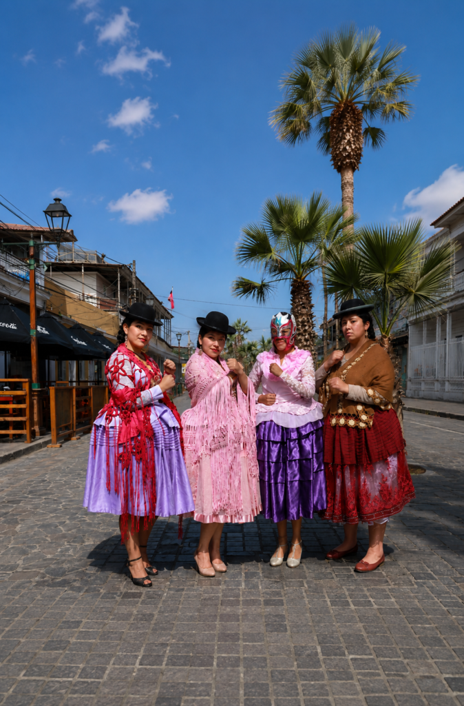 Cuatro cholitas luchadoras bolivianas posando juntas en el Paseo Baquedano de Iquique, Chile. Visten trajes tradicionales de pollera color violeta, rosa y rojo, con mantas bordadas, sombreros bombín negros y una de ellas lleva una máscara rosa de lucha libre mexicana.
