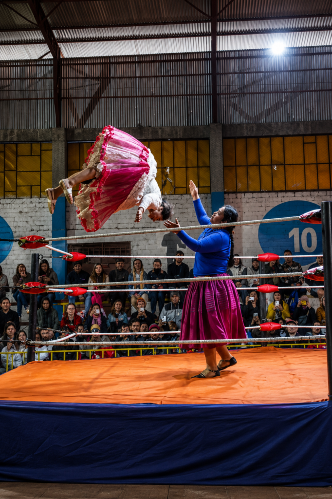 Una cholita luchadora boliviana realiza un salto acrobático invertido en el aire vistiendo una pollera fucsia con enaguas blancas. Abajo en el ring de lona naranja, otra luchadora con pollera morada y blusa azul estira los brazos en el coliseo de la zona 12 de Octubre en El Alto, Bolivia.