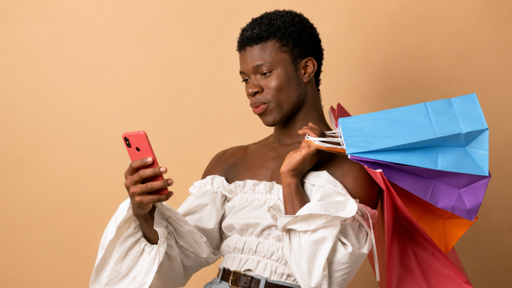 Una persona negra transgénero sonríe mientras interactúa con su teléfono inteligente sosteniendo bolsas de compras de colores sobre un fondo beige, representando las compras de moda en línea.