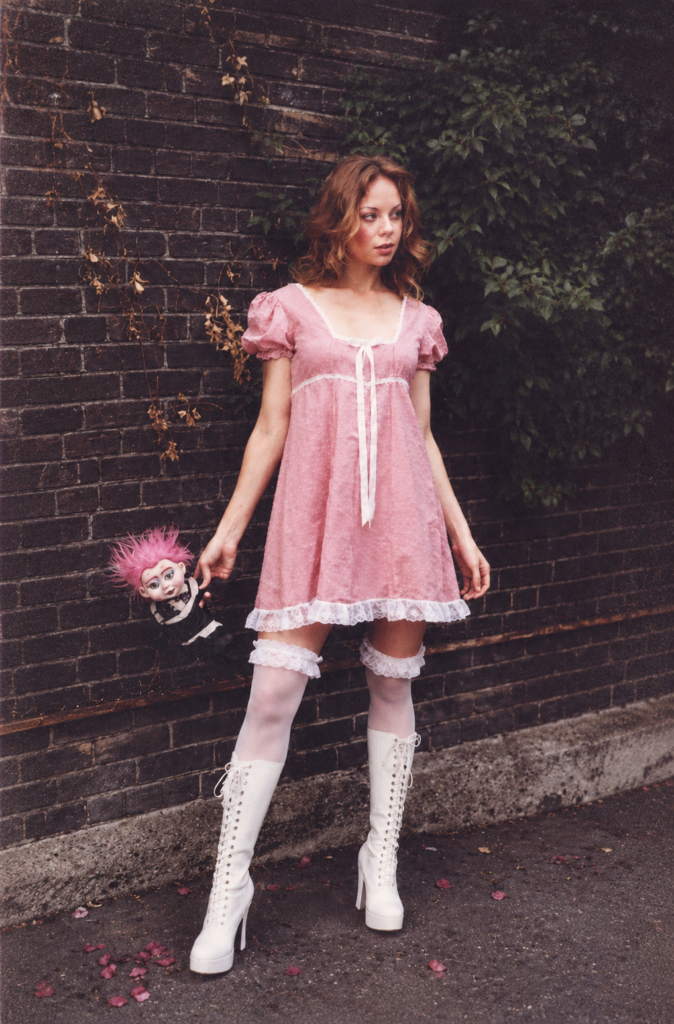 Una mujer rubia posa en la calle sosteniendo una muñeca con pelo rosa, vistiendo un minivestido babydoll rosa con encaje blanco, medias altas y botas blancas de plataforma.