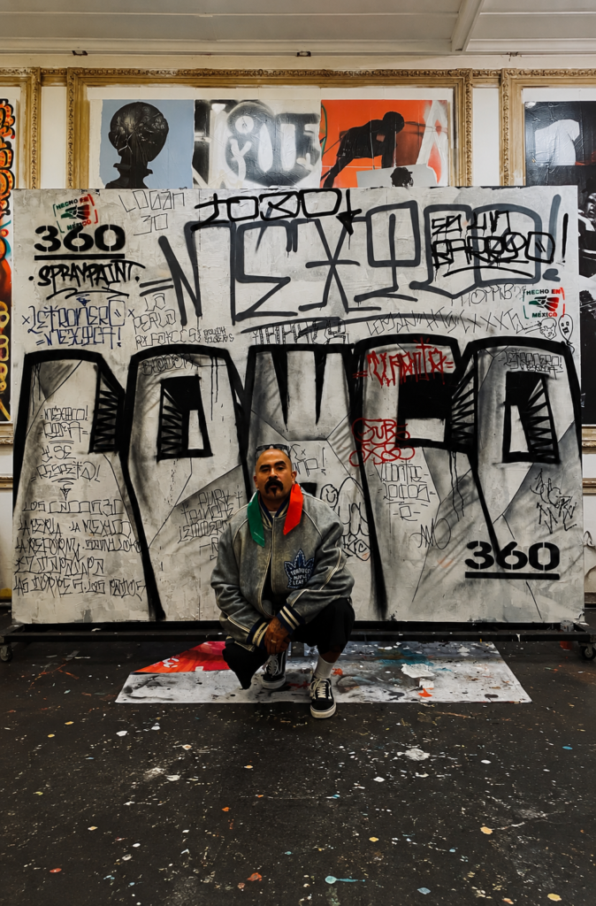 Marco Osorio de Letronero Mexicano posando con sombrero de lado y sosteniendo la bandera de México frente a un pilar pintado con un mural urbano de la firma yukarte77 en la Ciudad de México