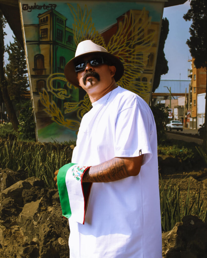 Marco Osorio de Letronero Mexicano posando con sombrero de lado y sosteniendo la bandera de México frente a un pilar pintado con un mural urbano de la firma yukarte77 en la Ciudad de México