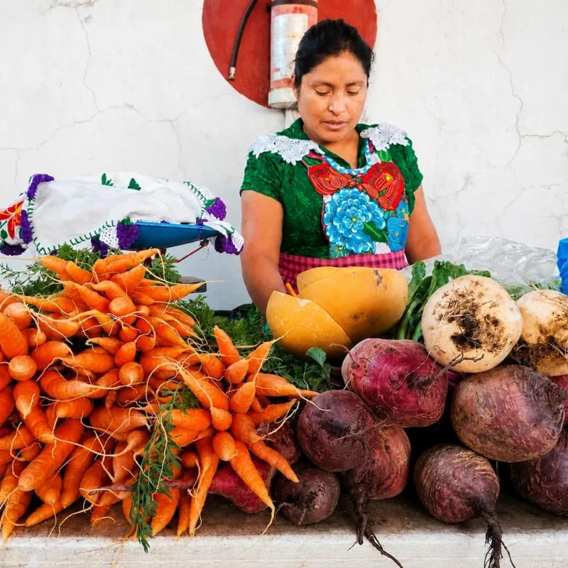 Mujer productora atendiendo un puesto con zanahorias frescas, betabeles y verduras de temporada en un mercado agroecológico de la Ciudad de México.