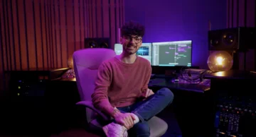 How independent musicians use AISong's Song Maker App Músico productor independiente sonriendo en su estudio de grabación casero, iluminado con luces LED moradas y azules, con equipo de audio profesional y pantallas mostrando software de producción musical DAW de fondo.