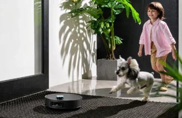 ¿Qué robot aspirador funciona mejor en hogares desordenados con limpiezas irregulares? Un robot aspirador de color negro está en primer plano sobre una alfombra texturizada oscura. A la derecha, un niño con camisa rosa y pantalones cortos corre felizmente. A su lado, un perro de pelaje gris y blanco salta en el aire. El fondo muestra una sala de estar con una gran planta de interior en una maceta cuadrada gris y fuertes sombras proyectadas sobre una pared blanca.