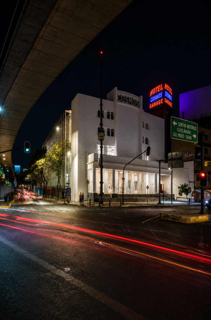 Vista nocturna vertical de la fachada blanca Art Decó restaurada del FARO Cosmos, antiguo Cine Cosmos, con estelas de luz de autos en la Calzada México-Tacuba, Ciudad de México.