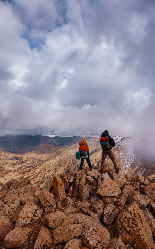 Dos excursionistas con mochilas de montaña contemplando un paisaje de cumbres rocosas y nubes densas desde la cima.