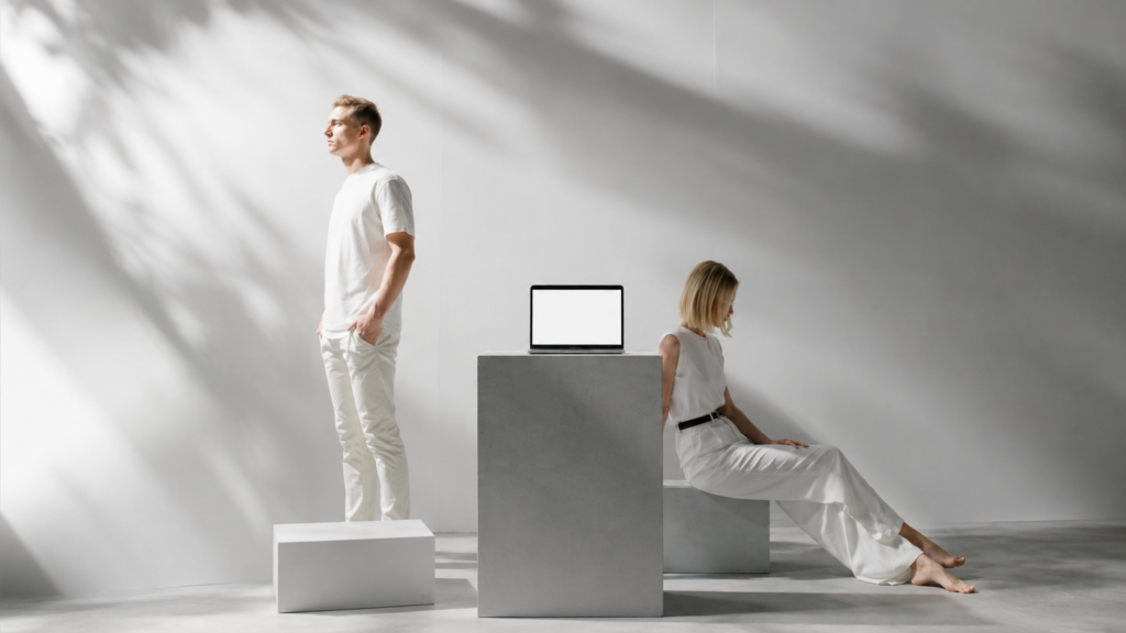 Un hombre de pie y una mujer sentada en un set de estudio minimalista completamente blanco con sombras de hojas, modelando ropa monocromática junto a una laptop sobre un pedestal para un catálogo de moda digital.