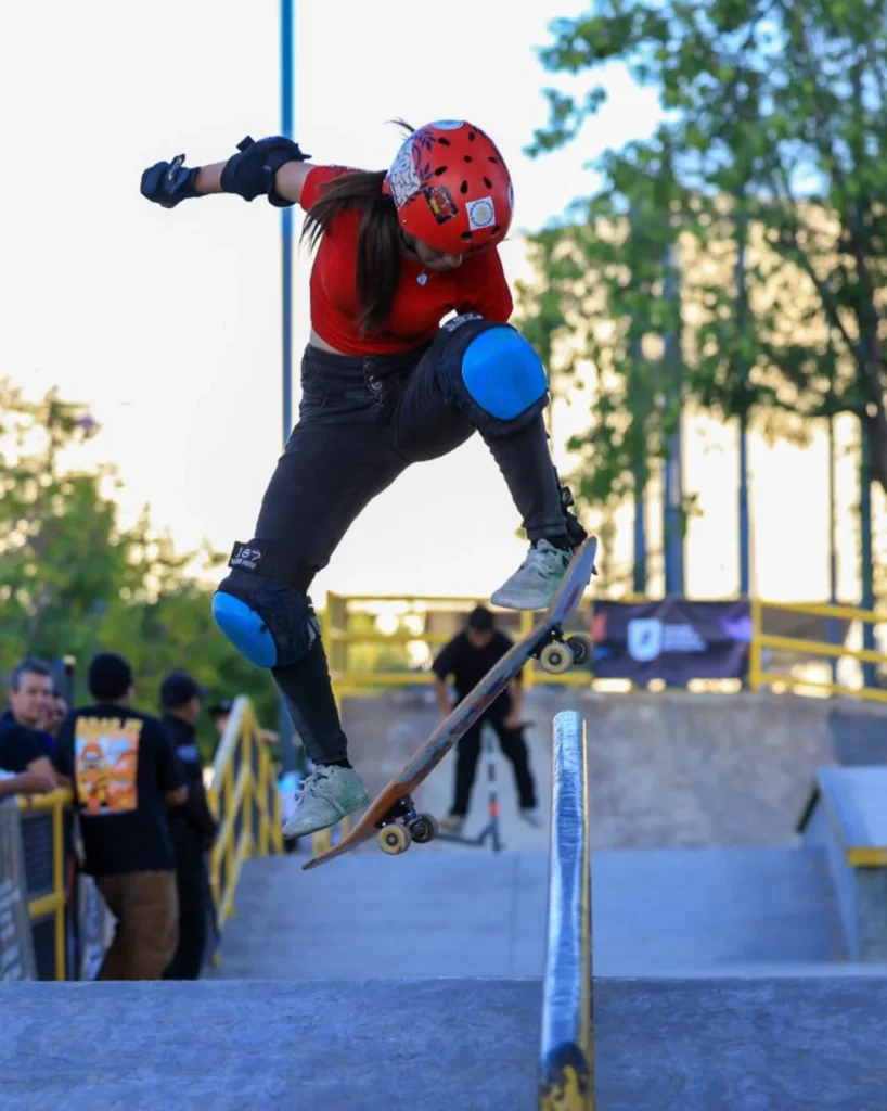 Una skater mexicana con casco rojo y rodilleras azules realiza un truco aéreo saltando sobre un largo barandal metálico en un skatepark.