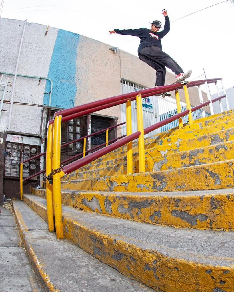 Un skater mexicano realiza un truco técnico deslizándose por un largo barandal rojo sobre unas escaleras amarillas de concreto en un spot de calle en México.